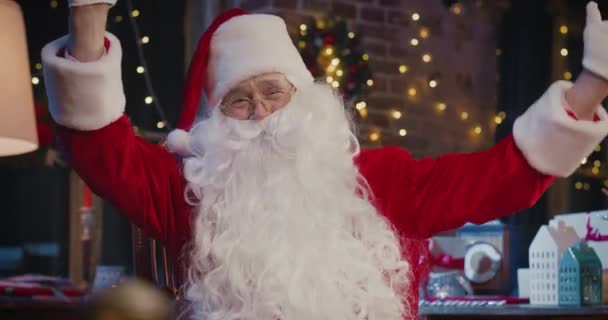 Święty Mikołaj siedzi przy fotelu i macha do kamery, siedząc w domu. Blisko szczęśliwego seniora w kostiumie Świętego Mikołaja. Koncepcja ducha Bożego Narodzenia — Wideo stockowe