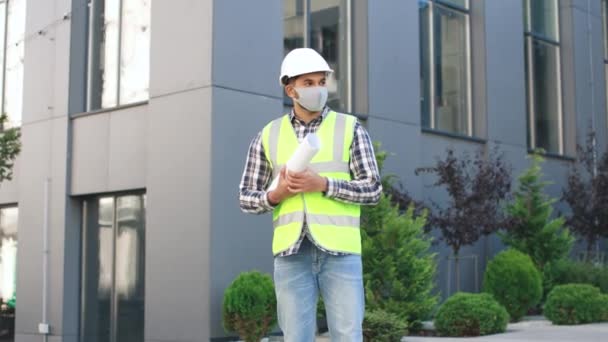 Dovedný kavkazský stavitel nebo architekt v ochranné masce a helmě stojící na ulici v blízkosti budovy a dívající se na kameru při pózování — Stock video