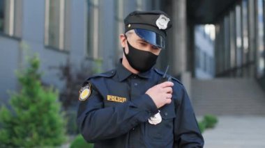 Koruyucu maske ve şapka takan yetenekli beyaz polis binanın yanında duruyor ve kameranın önünde radyoya konuşuyor.