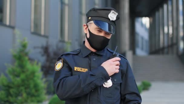Skillful caucasian policeman wearing protective mask and cap standing at the street near the building and speaks into the radio in front of the camera — Stock Video