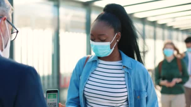 Κοντινό πλάνο του καυκάσιου ελεγκτή με μάσκα στο δρόμο έλεγχο της υγείας σάρωση QR κώδικα του εμβολιασμού ψηφιακό διαβατήριο σε εφαρμογή smartphone και τη μέτρηση της θερμοκρασίας του αφρο τουρίστα — Αρχείο Βίντεο