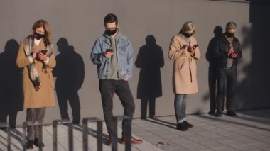 Güvenlik mesafesinde caddede durmuş, koruyucu maskeler takan ve aletlerine bakan insanların tam boy görüntüsü.