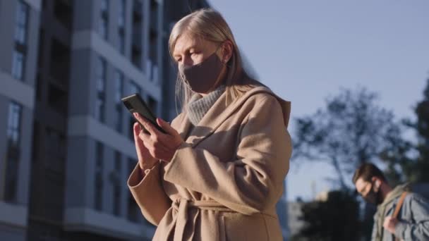 Vista verticale in vita dell'uomo e della donna che indossano maschere protettive in piedi sulla strada a distanza di sicurezza e che guardano i loro gadget — Video Stock