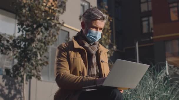 Cintura para cima vista retrato do homem sênior usando máscara protetora sentado no banco e trabalhando remotamente durante a pandemia. Novo conceito normal — Vídeo de Stock