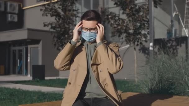Porträt des kaukasisch-kranken jungen Mannes mit Schutzmaske, der sich auf der Straße während einer Pandemie an seinem Kaugummi reibt und Kopfschmerzen verspürt — Stockvideo
