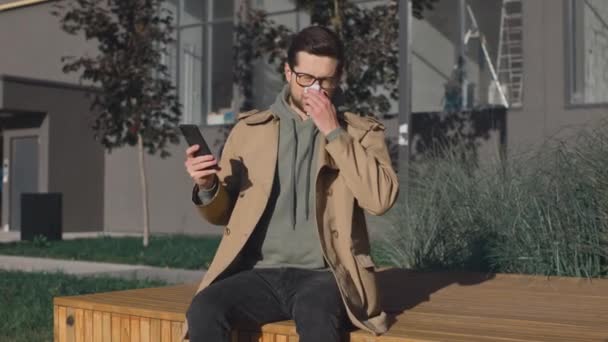 Sick caucasian man sneezing on city street background. Ill male sad guy with cold symptoms sneeze outdoors while using smartphone at the bench. Portrait of the male suffering from allergy and feeling — Stock Video