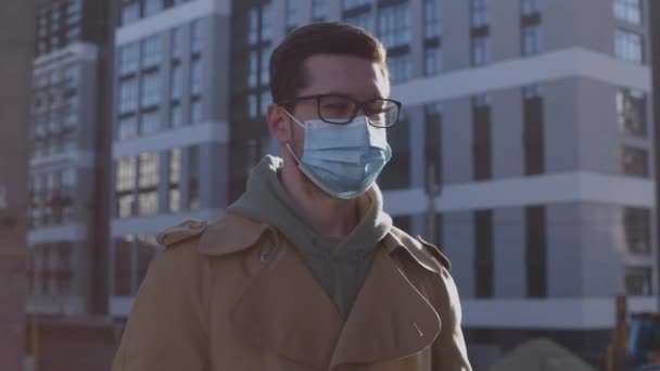 Derékig érő portré a fehér fiatalemberről, aki védőmaszkot visel, magabiztosan pózol az utcán a járvány idején. — Stock videók