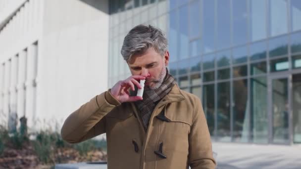 Senior man wearing plaid scarf spraying nasal spray while standing in the city street. Health care therapy and medicine concept — Stock Video