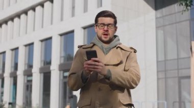 Kafkasyalı hasta bir adam şehrin arka planında hapşırıyor. Soğuk algınlığı belirtileri olan hasta bir erkek akıllı telefon kullanırken dışarıda hapşırıyor. Alerjisi olan ve kötü hisseden bir erkeğin portresi.