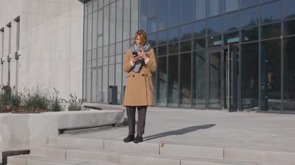 Vol lngth van de Kaukasische vrouw hoesten hard op straat in de herfst met behulp van haar smartphone. Kantoorgebouw achter haar — Stockvideo