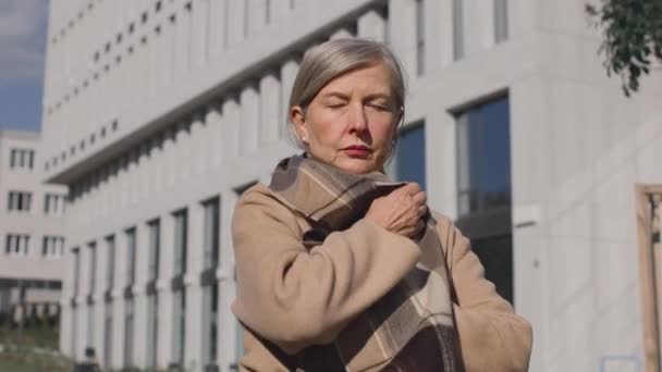 Egy idős, fagyott nő meleg kabátban. Gyönyörű nő becsomagolva a ruhájába, miközben sóvárog a hideg állva az őszi utcában. A felmelegedés fogalma — Stock videók