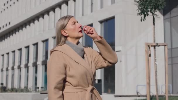 Mujer mayor con bufanda gris rociando aerosol nasal mientras está de pie en la calle de la ciudad. Terapia sanitaria y concepto de medicina — Vídeos de Stock