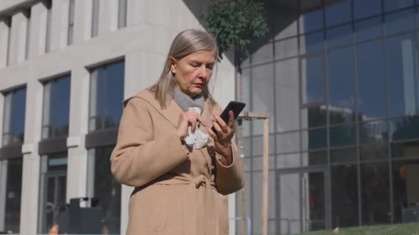 Kranke Kaukasierin, die auf der Straße niest. Kranke Frauen mit Erkältungssymptomen niesen im Freien, während sie ihr Smartphone benutzen. Porträt der Frau, die an Allergie leidet und sich schlecht fühlt — Stockvideo