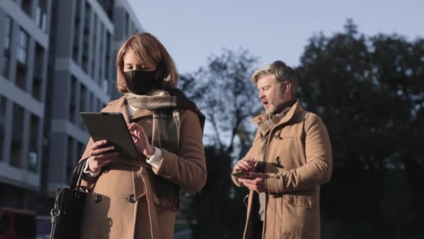 Distância de segurança. Cintura para cima vista retrato do homem e da mulher vestindo máscaras protetoras em pé na rua e sendo profundo nos gadgets durante a pandemia covid 19 — Vídeo de Stock