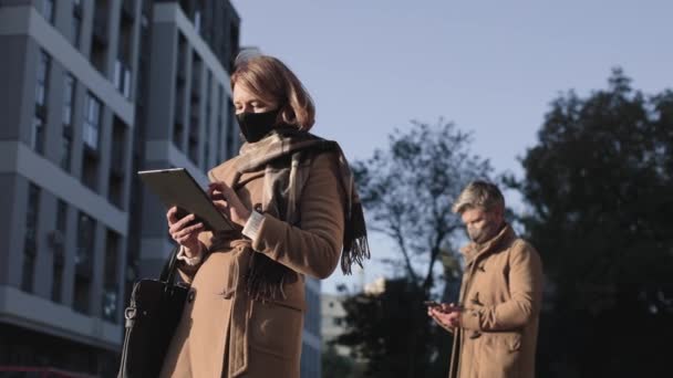 Cintura hacia arriba retrato vista del hombre y la mujer con máscaras protectoras de pie en la calle a la distancia de seguridad y mirando sus aparatos — Vídeos de Stock