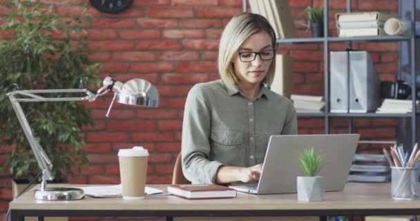 Portræt af smuk og succesfuld blondine forretningskvinde med briller, der arbejder i moderne kontor ved hjælp af bærbar computer og smilende ser på kameraet. – Stock-video