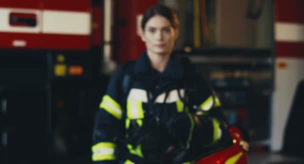 Camera zooming in the good looking young Caucasian woman in the special firefighter uniform and with a helmet in a hand standing and smiling on the fire truck background. From blurred to focus — 图库视频影像