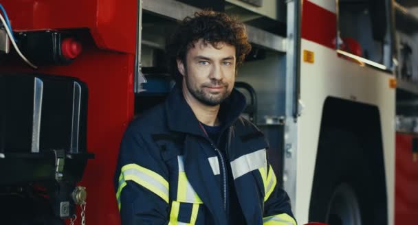Portrét kavkazského dobře vypadajícího hasiče v plně vybavených šatech, jak se dívá do strany a pak se otočí tváří ke kameře u červeného hasičského vozu. — Stock video