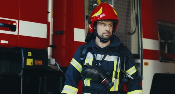 Portrait du beau garde-feu caucasien en costume équipé et casque regardant sur le côté, puis tournant la tête vers la caméra sur le fond rouge du camion de pompiers. — Video