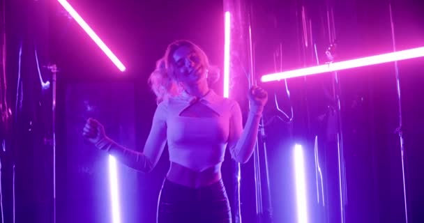 Junge kaukasische Frau mit lockigem Haar tanzt im Nachtclub zwischen Neonlichtern. Fröhliche blonde Mädchen in engen Top und Lederhosen lächeln und haben Spaß allein auf der Party. — Stockvideo