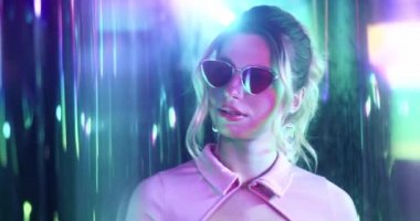 Kıvırcık saçlı ve güneş gözlüklü genç beyaz kadın renkli ışıkların arasında duruyor, yukarı bakıyor. Gece kulübünde merak eden çekici sarışın kıza yaklaş..