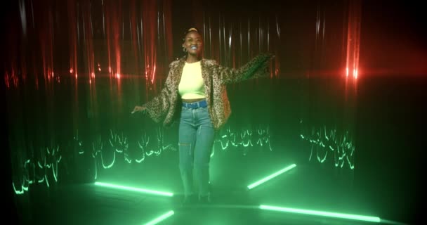 Wanita muda Afrika-Amerika dengan celana jeans biru, mantel bulu dan kacamata hitam menari di antara lampu neon. Happy girl menikmati musik, bersenang-senang sendirian di pesta di lampu hijau klub malam. — Stok Video