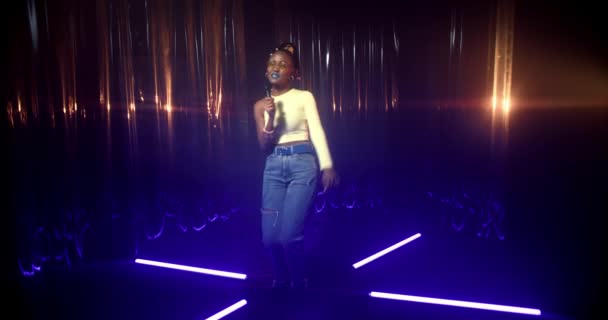 Joven mujer afroamericana en pantalones vaqueros azules y flaco top baila y canta en el micrófono entre luces azules de colores. Chica feliz gestos y disfruta de la música, realizando en la fiesta en el club nocturno. — Vídeos de Stock