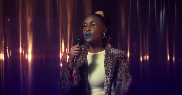 Joven mujer afroamericana en abrigo de piel y gafas de sol baila y canta en el micrófono entre luces de colores. Chica feliz gestos y disfruta de la música, realizando en la fiesta en el club nocturno. — Vídeos de Stock