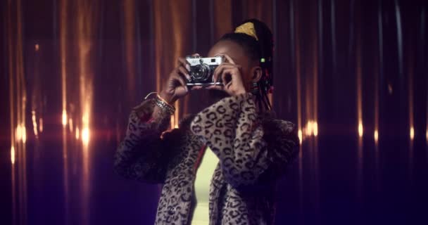 Young African American woman in fur coat and skinny top makes photos with film camera, then smiles. Happy girl in party outfit with bold makeup taking pictures. — Stock Video