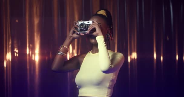 Junge Afroamerikanerin in dünnem Top macht Foto mit Filmkamera, dann lächelt sie. Glückliches Mädchen im Party-Outfit mit fettem Make-up beim Fotografieren. — Stockvideo