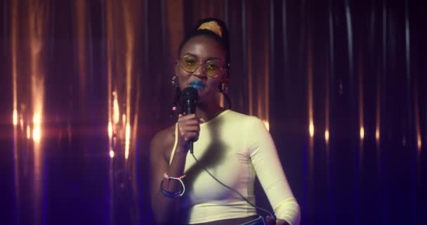 Giovane donna afroamericana in magro top danza e canta in microfono tra luci blu e gialle. Ragazza felice con gesti di trucco audaci e gode di musica, esibendosi alla festa nel night club. — Video Stock