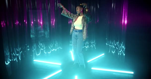 Wanita muda Afrika-Amerika dengan celana jeans biru, mantel bulu, dan baju dansa tipis di antara lampu neon. Happy girl menikmati musik, bersenang-senang sendirian di pesta di lampu pirus klub malam. — Stok Video