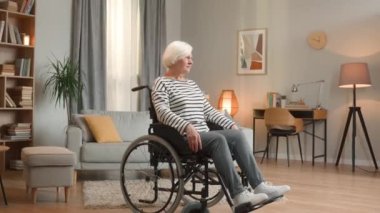 Oturma odasında tekerlekli sandalyede tek başına oturan son sınıf öğrencisi kadın kameraya bakıyor. Beyaz saçlı kadın, ev içinde, rahat bir dairede özürlü. Hastalık, evde kameraya gülümseyen yalnız insan.
