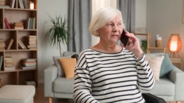 Engelli, güzel, yaşlı bir kadın emeklinin rahat oturma odasında akıllı telefondan konuşmasını kapatın. Gri saçlı mutlu bir kadın cep telefonundan arıyor ve boş zamanlarında sohbet ediyor.