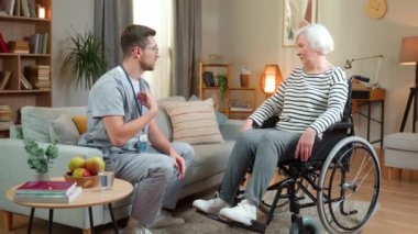 Yakışıklı genç erkek doktor oturma odasında tekerlekli sandalyedeki engelli kadın hastayla konuşuyor. Erkek rehabilitasyon terapisti yaşlı kadını evde ziyaret etti, tıbbi ve sağlık tedavisi kavramı