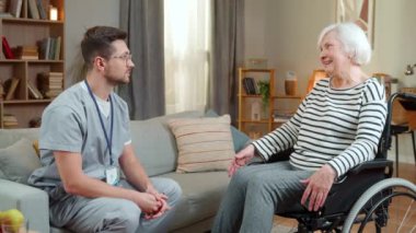Tekerlekli sandalyede oturan engelli bir kadınla konuşan genç bir erkek doktorun yan görüntüsü. Sağlık çalışanı evde kıdemli bir hastayı ziyaret ediyor ve onunla konuşuyor, rehabilitasyon konsepti