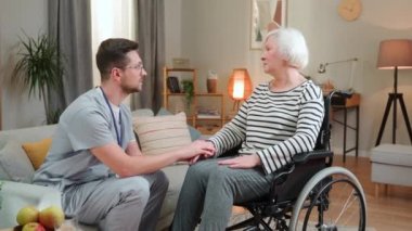 Üniformalı yakışıklı genç adam tekerlekli sandalyedeki yaşlı kadın hastayla konuşuyor, psikolog, rehabilitasyon, destek. Erkek hemşire evde yaşlı bayanla konuşuyor ve kameraya gülümsüyor.