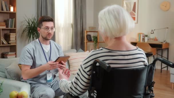 男医生 年轻的专业男护士在与老年残疾妇女在公寓 家庭护理 保健概念等方面交谈时 在平板电脑上敲了一下 — 图库视频影像