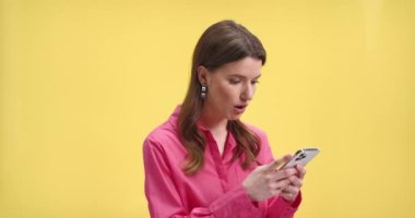 Stüdyoda sarı arka planda duran pembe bluzlu neşeli genç bir kadının sosyal ağ uygulamasında akıllı telefonuna şaşkın yüz ifadesiyle göz atarken yakın plan portresi.