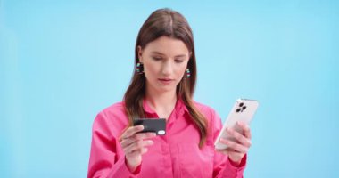 İnternet üzerinden alışveriş yapan mutlu genç bir kadının portresi. Cep telefonuyla mavi arka planda duruyor ve gülümsüyor. Evet hareketi yapıyor. Cep telefonuyla kadın alışverişi, kredi kartıyla ödeme, kapat.