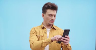 İnternet üzerinden alışveriş yapan mutlu bir gencin portresi cep telefonuyla mavi arka planda duruyor ve gülümsüyor. Evet hareketi yapıyor. Cep telefonuyla erkek alışverişi, kredi kartıyla ödeme, kapat.