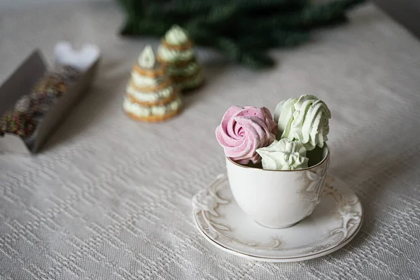 Pembe ve yeşil büyük marşmelovlar şekerleme ağaçlarının ve ladin dallarının arka planında bir fincanda uzanır.