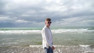 Beyaz gömlekli genç bir adam deniz boyunca mutlu bir şekilde yürür.