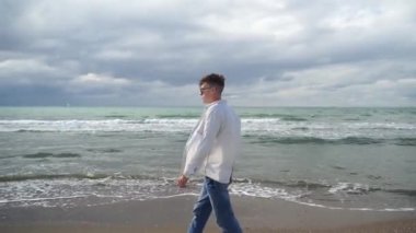 Tişörtlü modern genç adam kumlu deniz kıyısında yürüyor, güneşli bir günde.