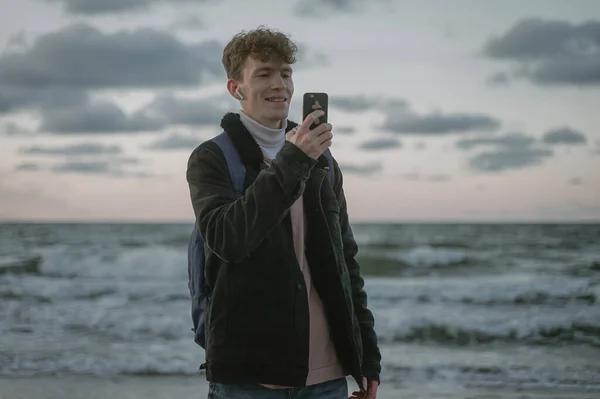 Kıvırcık saçlı, gülümseyen, cep telefonuyla fotoğraf çeken, elinde akıllı telefon tutan, Z neslinden mutlu genç bir adam.