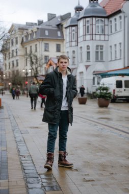 Genç bir adam sokağın ortasında duruyor, ellerini cebinde tutuyor, ciddi bir yüzle kameraya bakıyor, siyah kışlık ceketli, soğuk, nemli bir şehir.
