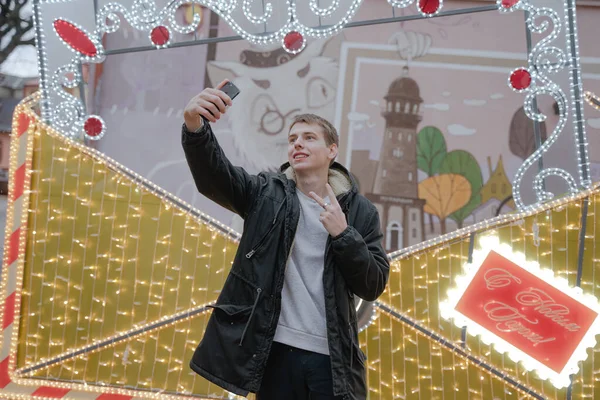 Adam poz veriyor, sosyal ağlar için fotoğraf çekiyor, yılbaşı atmosferi, iki parmağını gösteriyor, telefonu elinde tutuyor. Telefonda selfie, kameraya gülümsüyor.