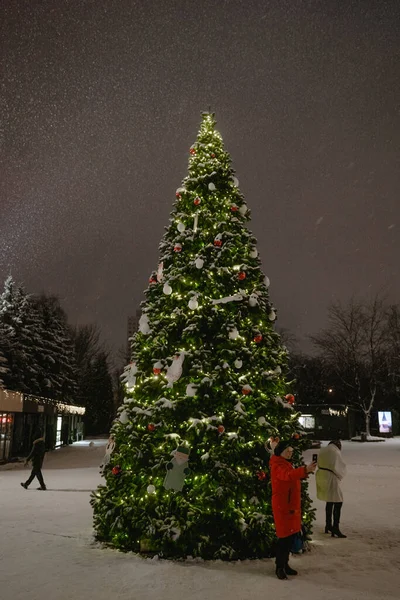 İnsanlar gece yılbaşı ağacında fotoğraf çekiliyor. Gece sokağa kar yağıyor, Noel ışıkları yanıyor.