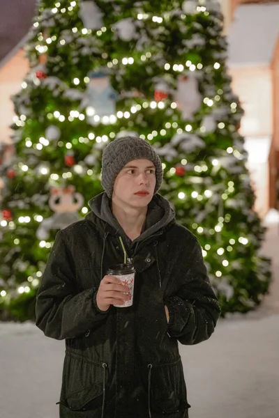 Soğuk kış mevsiminde Noel ağacının yanında sıcak bir içecek içer. Şehir ortamında kışlık giysiler giyer.
