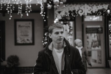 Kışlık ceketli bir genç, yılbaşı ışıklarının arka planında, yana bakıyor, siyah ve beyaz genç adamı şımartıyor.